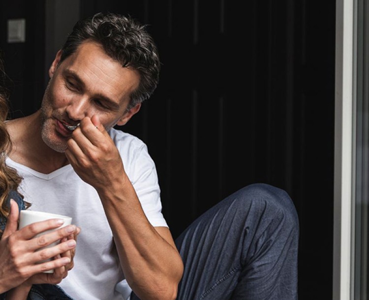 Twee personen zitten lachend en praten bij een open deur, de vrouw houdt een kopje vast.