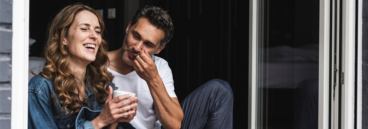 Twee personen zitten lachend en praten bij een open deur, de vrouw houdt een kopje vast.