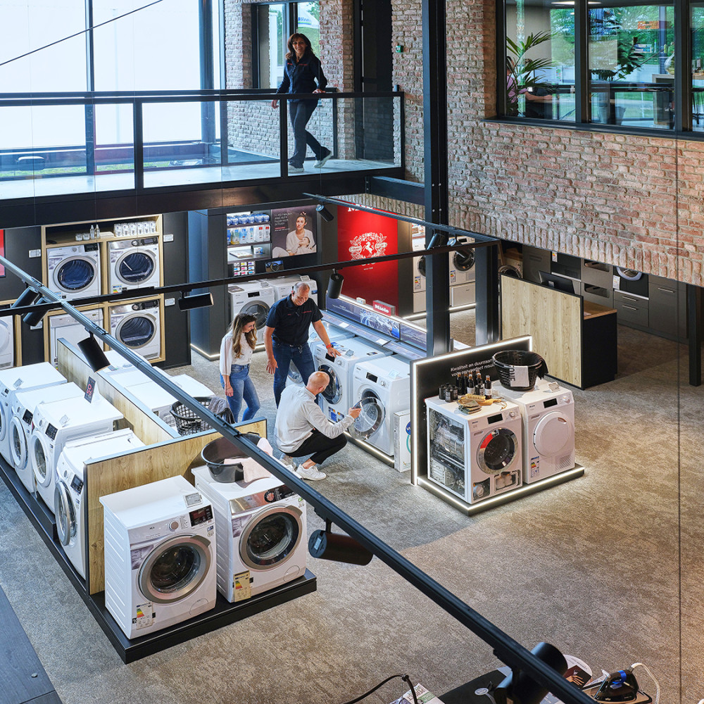 Winkel met mensen die naar wasmachines en drogers kijken; modern interieur met glazen balustrade.