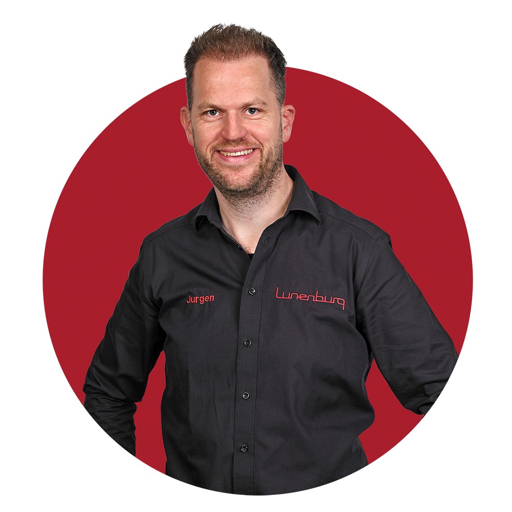 Man in een zwart overhemd met de naam "Jurgen" erop geborduurd, voor een rode cirkelachtergrond.