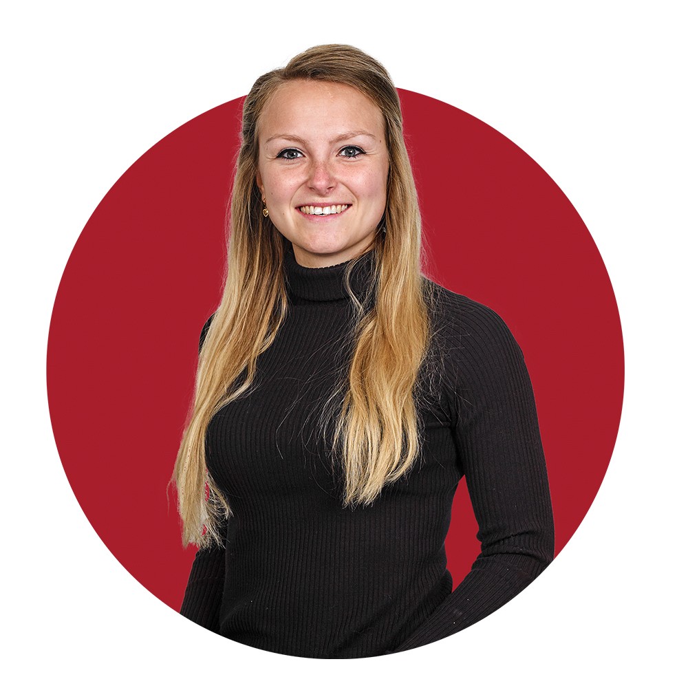 Persoon met lang blond haar en zwarte trui poseert voor een rode cirkelvormige achtergrond.