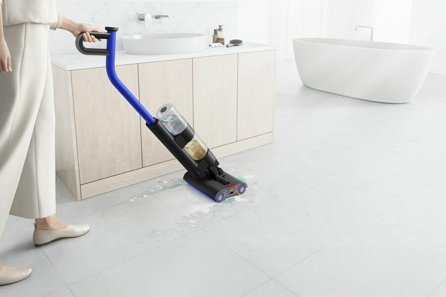 Persoon gebruikt een elektronische dweil op een tegelvloer in een badkamer met een bad en wastafel.