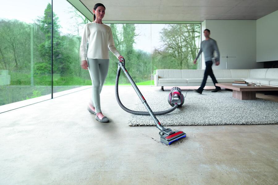 Een vrouw stofzuigt een moderne woonkamer met een grote glazen wand en zicht op de tuin.