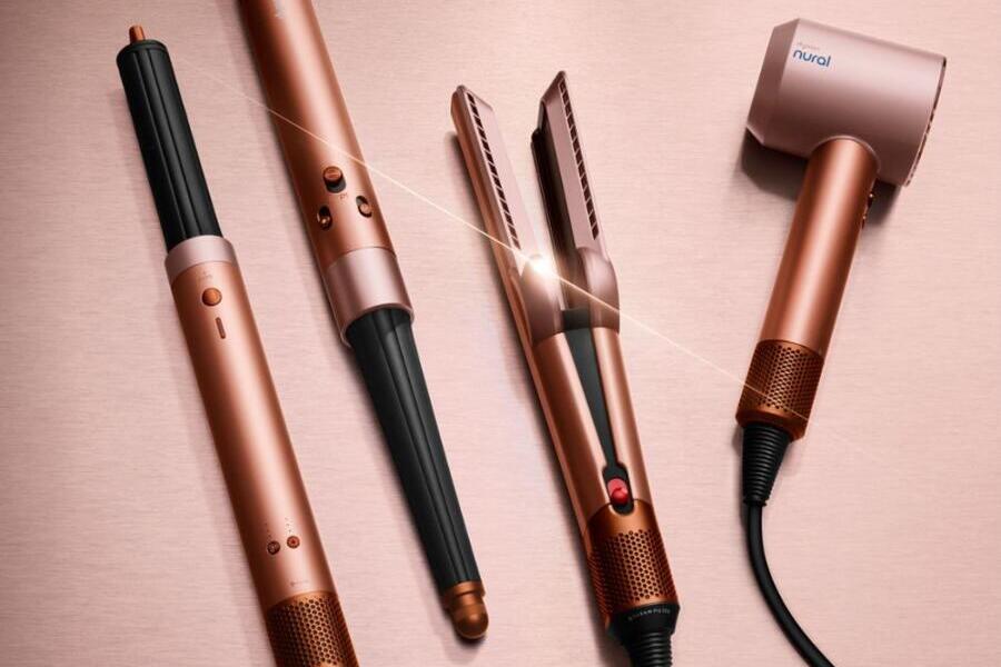 Roze en koperkleurige haarstylingtools: krultangen, stijltang en föhn op houten achtergrond.